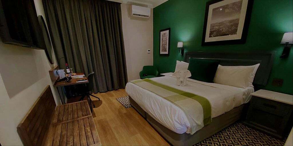 A spacious hotel guest room featuring a king-size bed with a green accent wall, a wooden work desk, and a seating area at Indaba Hotel Spa and Conference Centre in Johannesburg.