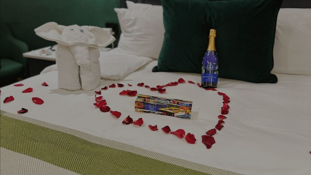 A neatly made hotel bed decorated with a heart-shaped arrangement of red rose petals, a bottle of sparkling wine, and an origami towel elephant at Indaba Hotel Spa and Conference Centre in Johannesburg.