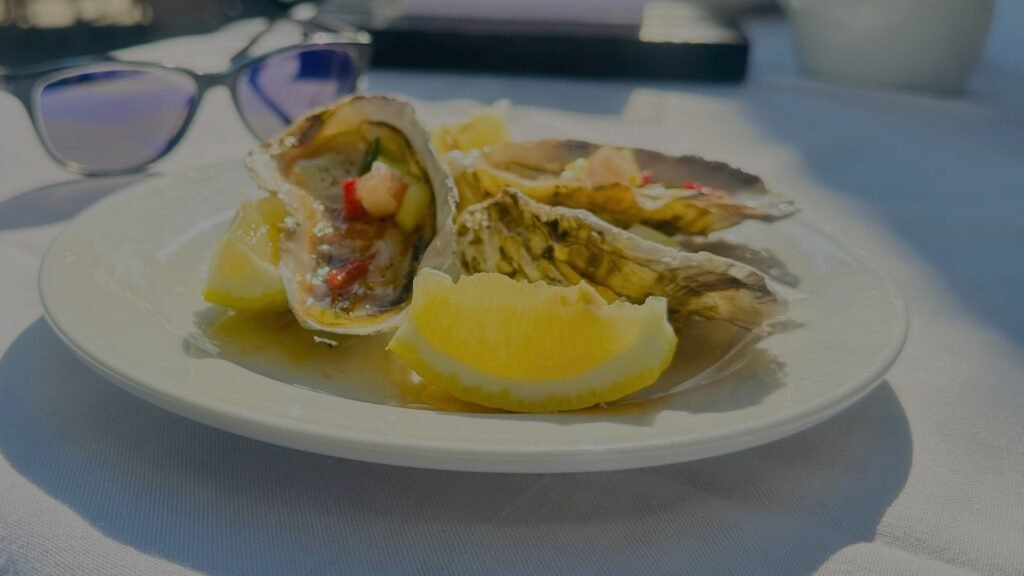 Freshly shucked oysters garnished with seasoning and fresh lemon wedges on a white plate next to a pair of sunglasses.