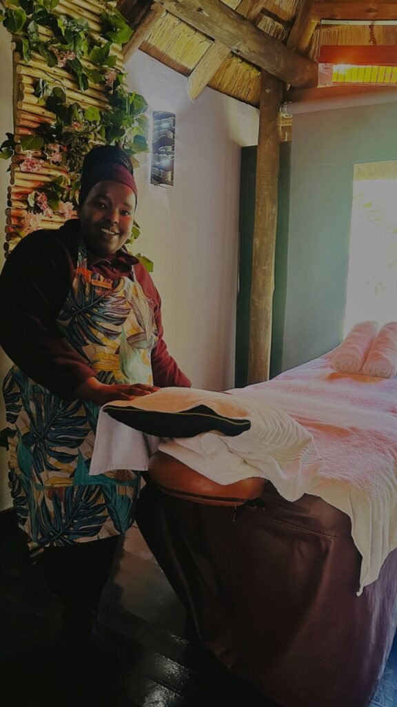 A smiling spa therapist wearing a floral apron standing next to a prepared massage table with clean towels at Indaba Hotel Spa and Conference Centre in Johannesburg.