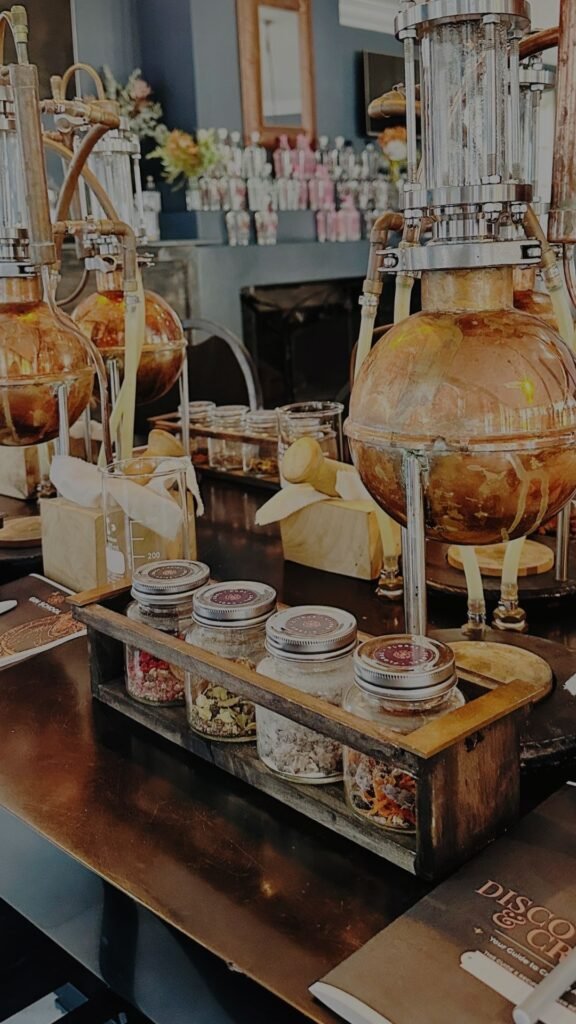 A detailed view of copper gin stills paired with a wooden caddy filled with glass jars of colorful botanicals on a dark tabletop.
