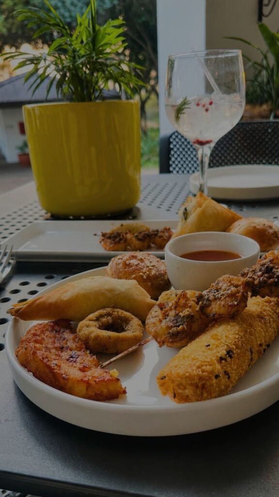 An assortment of savory pastries, skewered treats, and a dipping bowl on a white plate, served alongside a garnished cocktail glass and a yellow potted plant.