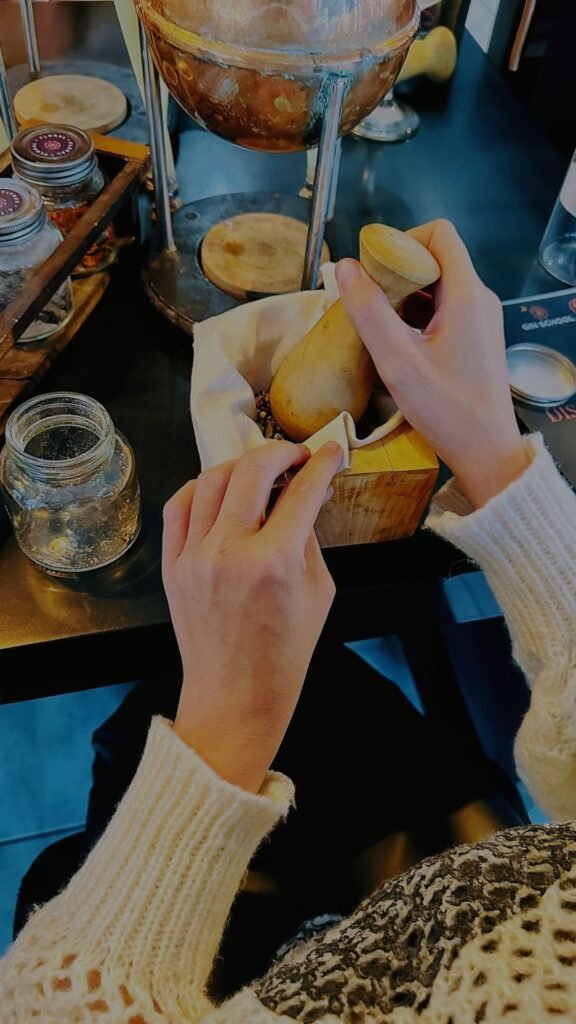 A close-up of hands using a wooden pestle and mortar to crush ingredients over a linen cloth, surrounded by botanical jars and distillation gear.