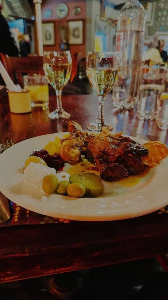 A white dinner plate piled high with roasted meats, poultry, olives, pickles, and side dishes on a wooden table at Indaba Hotel Spa and Conference Centre in Johannesburg.