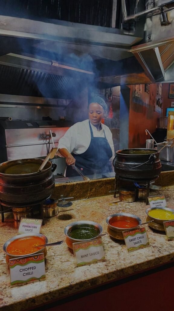 A chef in a white chef coat and black apron working in a restaurant kitchen behind a counter featuring bowls of various sauces at Indaba Hotel Spa and Conference Centre in Johannesburg.