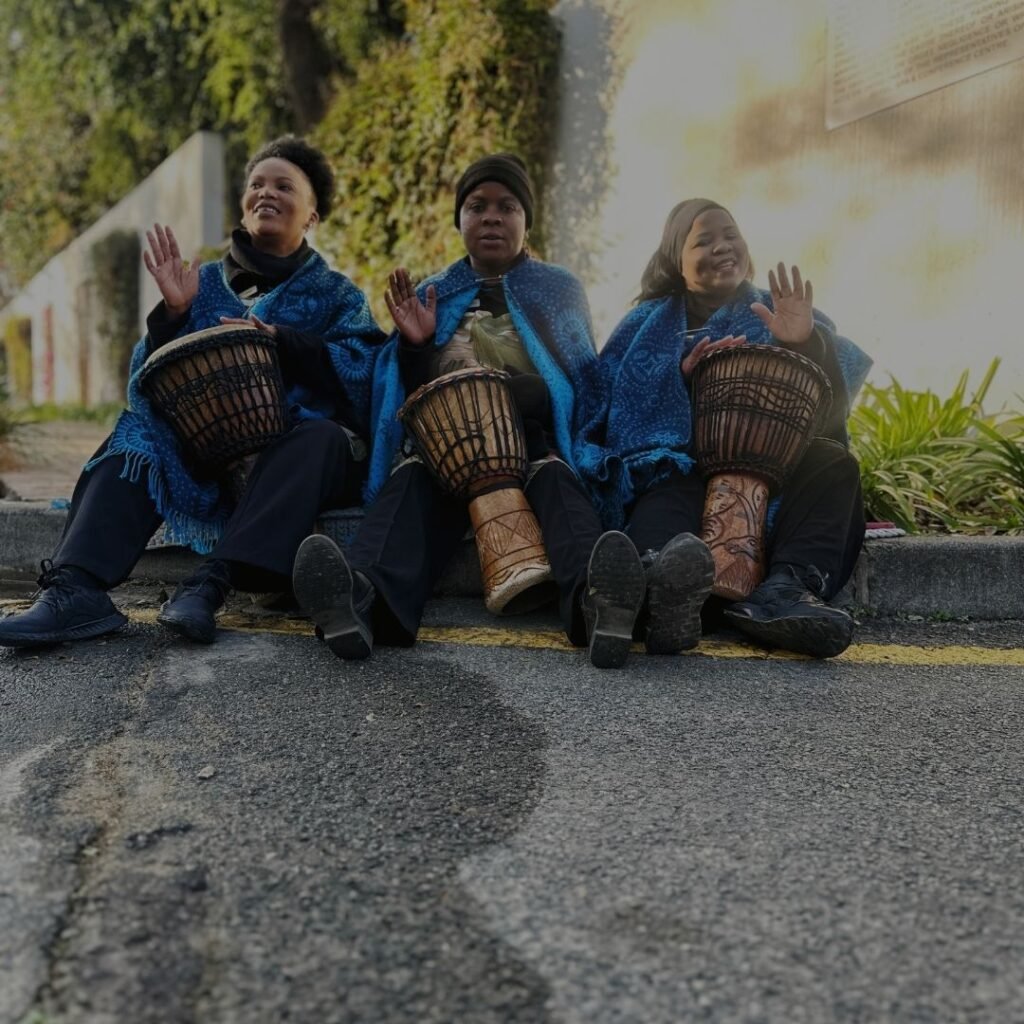 Three smiling women wearing traditional blue wraps and playing djembe drums while sitting on a curb at Indaba Hotel Spa and Conference Centre in Johannesburg.