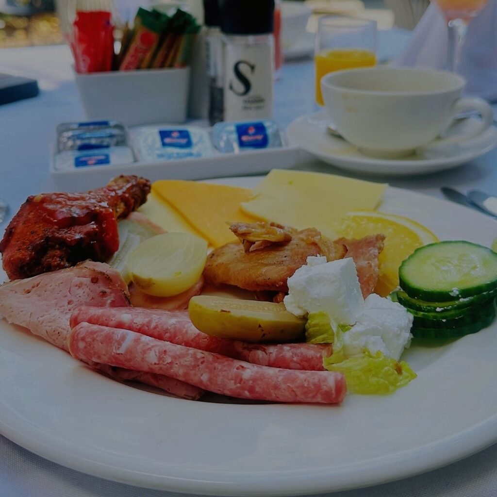 A white plate featuring a selection of cold meats, sliced cheeses, cured sausages, cucumber, and feta cheese alongside a coffee cup and butter packets at Indaba Hotel Spa and Conference Centre in Johannesburg.