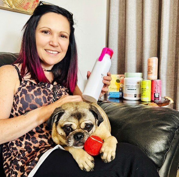 Claudia Sherrin smiling while holding a Beauty Gen collagen bottle next to her pug, with Naked Collagen Complete and iced teas in the background