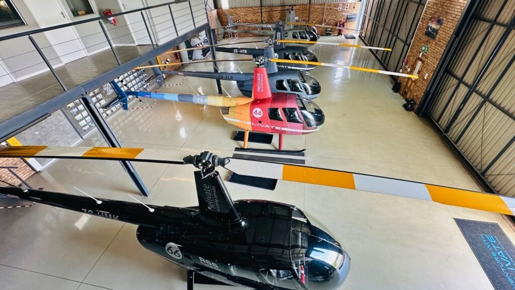 Multiple Helivate helicopters lined up inside the modern hangar at Jack Taylor Airport in Krugersdorp, including the black R66 Turbine ZS-HMK and the colourful Robinson R44 ZS-RYM number 44.