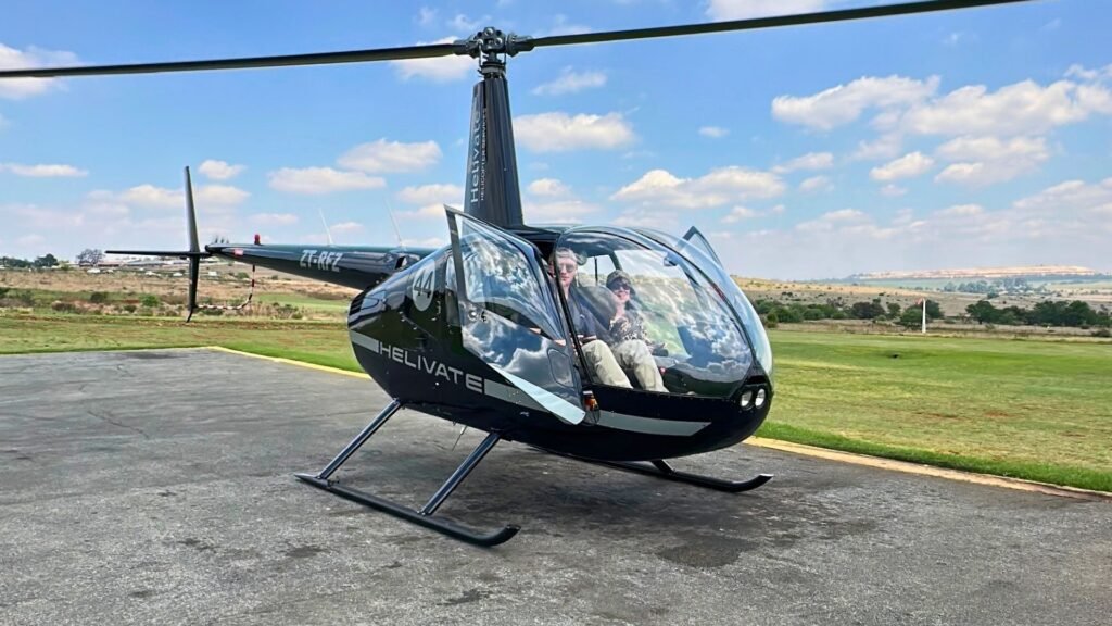 Claudia Sherrin and pilot Rory Root seated inside a black Helivate Robinson R44 helicopter (ZT-RFZ) with the door open on the tarmac under a clear blue sky.