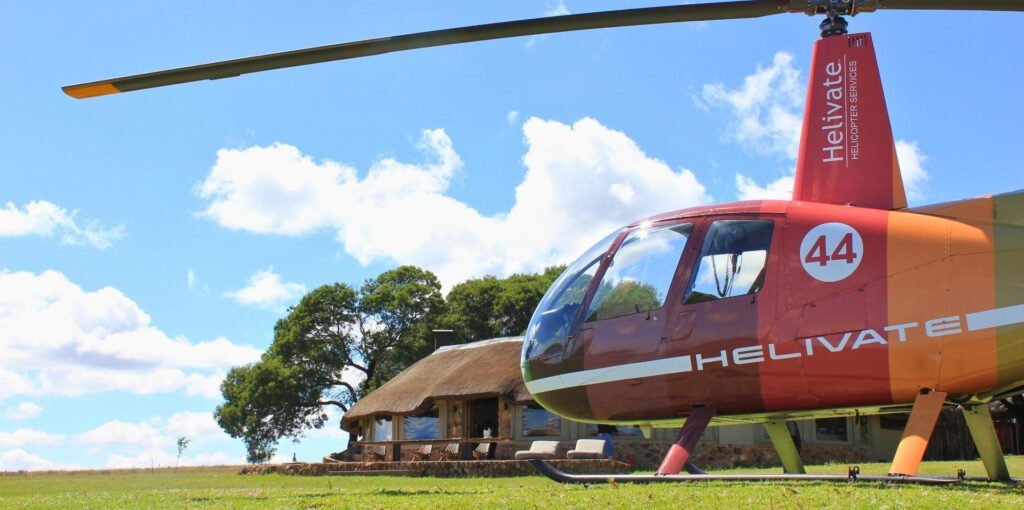 Colourful Helivate Robinson R44 helicopter number 44 parked on the grass in front of a thatched-roof lodge building under a bright blue sky with white clouds.
