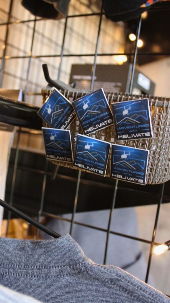 Square enamel pins reading “I Adventured with Helivate” featuring a helicopter and mountain graphic, displayed on a wire rack above branded apparel at Helivate Helicopter Services.