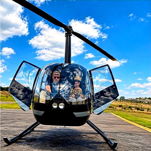 Claudia Sherrin and a male companion smiling from the open doors of a black Helivate helicopter on the tarmac under a clear blue sky.