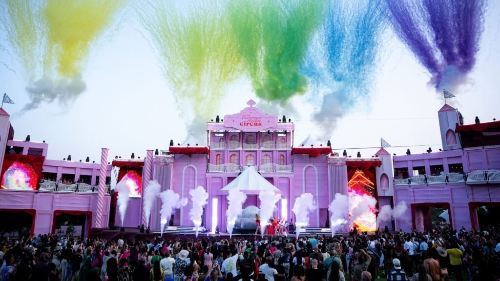 A wide view of the grand pink festival stage with vibrant bursts of colored smoke and pyrotechnics over a large crowd at The Luxurious Marble Circus.