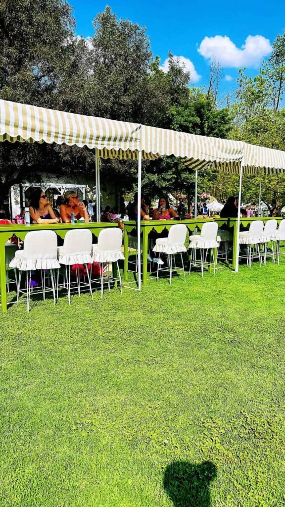 Guests seated along green tables under cream and taupe striped awnings on the lawns at the Luxurious Marble Circus.