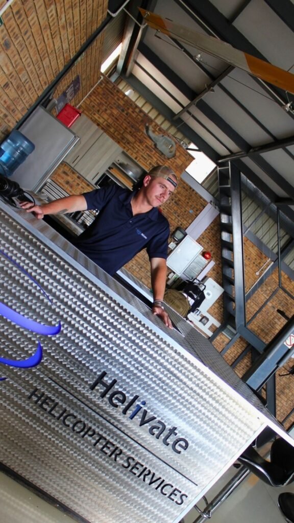 Rory Root in a navy polo and cap leaning on the branded metal reception counter inside the hangar at Jack Taylor Airport, Krugersdorp.
