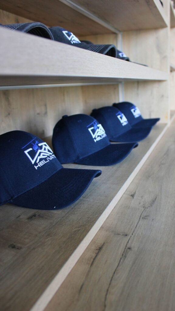 Row of navy blue Helivate baseball caps with white embroidered helicopter logo arranged on a light wooden shelf in the merchandise area.