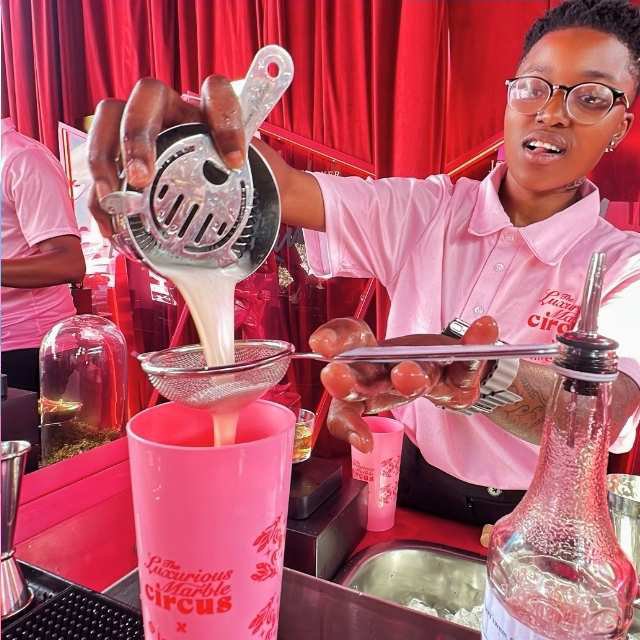 A bartender in a pink shirt straining a cocktail into a branded cup at the bar during the Luxurious Marble Circus event.