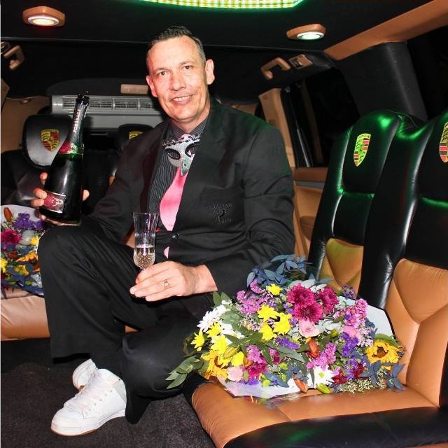 Judd Sherrin seated in the Porsche Cayenne limousine holding champagne and a colourful bouquet, smiling in formal attire
