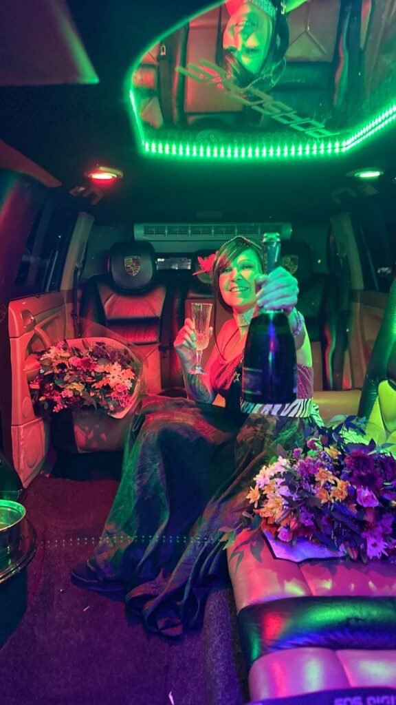 Claudia Sherrin smiling and raising a champagne bottle under green neon lights inside the Luxury Limousines Porsche Cayenne