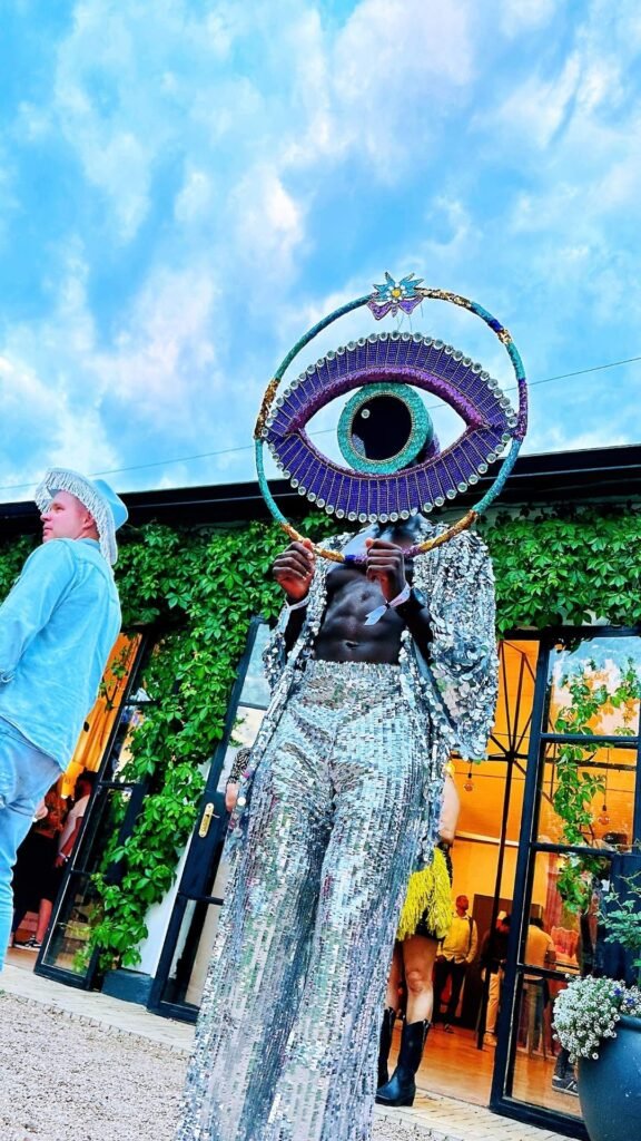 A performer in shimmering silver sequined attire holding a large decorative eye headdress outside a venue at the Luxurious Marble Circus.