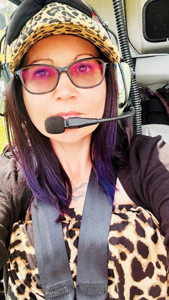 Claudia Sherrin wearing a leopard-print cap, pink-tinted sunglasses and aviation headset while seated inside a Helivate helicopter.