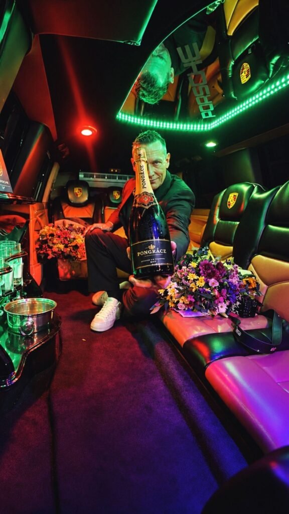 Judd Sherrin kneeling in the limousine holding a bottle of Pongrácz Méthode Cap Classique toward the camera amid flowers and LED lights