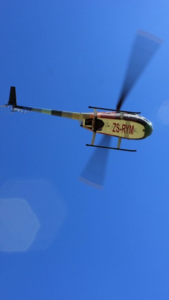 Colourful Helivate Robinson R44 helicopter registration ZS-RYM in flight against a clear deep blue sky with spinning rotors.
