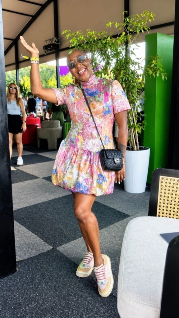 Mo smiling and waving in a vibrant floral dress and sunglasses indoors at the Luxurious Marble Circus venue.