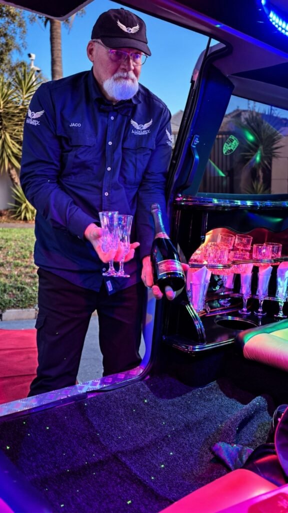 Chauffeur Jaco from Luxury Limousines holding crystal champagne flutes and a bottle at the open wing door of the white stretch limo